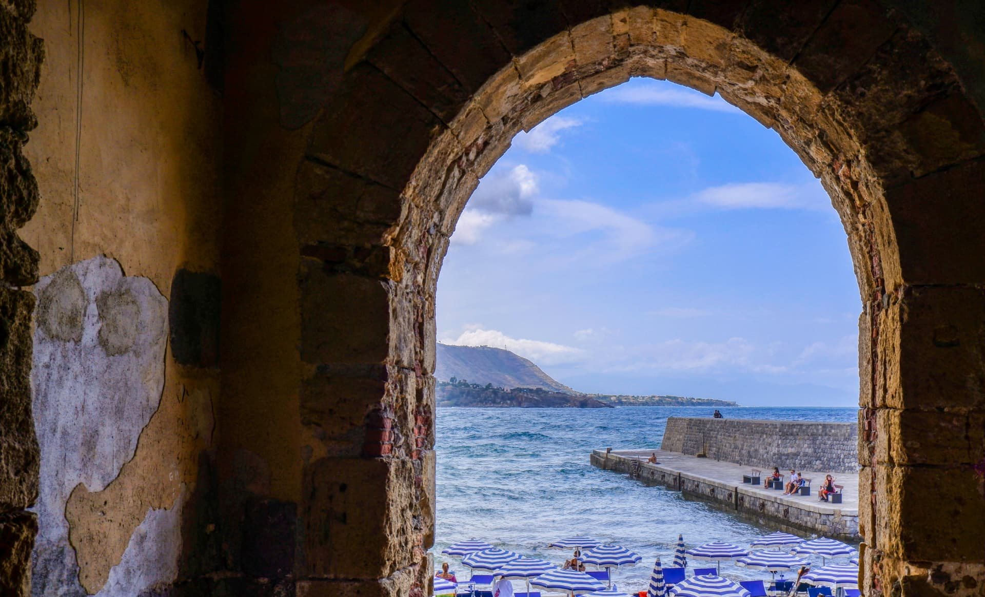 La spiaggia e il mare visti attraverso un arco di pietra, con gli ombrelloni e il molo